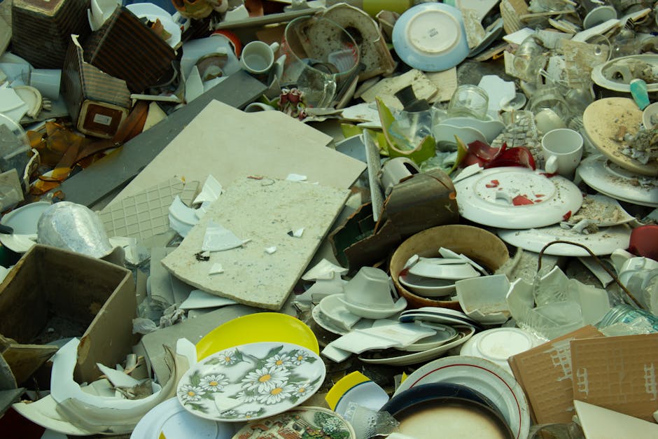 The image depicts a large pile of assorted discarded ceramics, porcelain, and glassware, including broken plates, cups, bowls, and vases, scattered across a hard surface such as a driveway or outdoor 