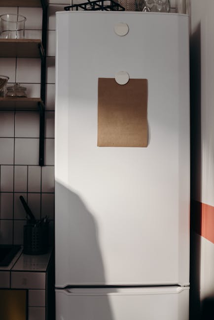 A tall, white, upright fridge freezer situated in a kitchen environment, with a brown paper note attached to the front door using round white magnets. The fridge has a smooth, glossy surface without v
